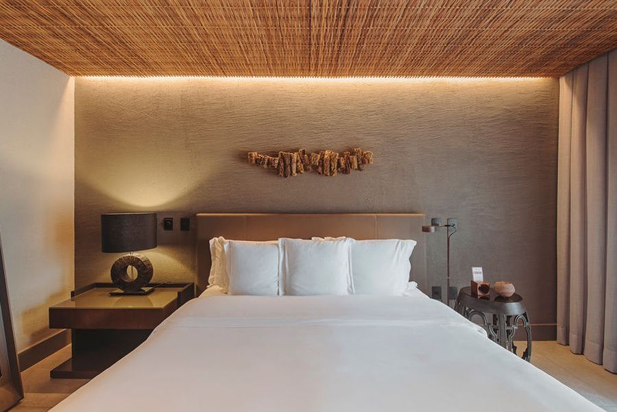 Um quarto de hotel moderno com cama branca, teto com detalhes em vime, parede marrom texturizada e mesas de cabeceira minimalistas com abajures.