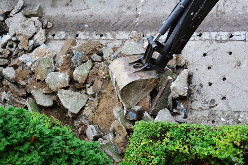 Excavator bucket demolishing a concrete wall, debris scattered near green bushes.