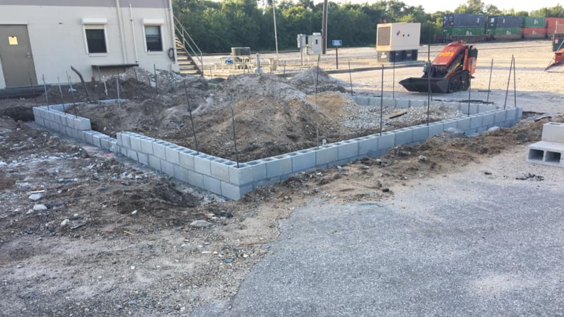 Construction site with concrete block foundation partially built; excavator in the background.