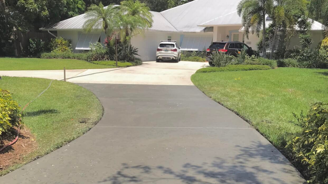 Curving gray driveway leads to a white house with cars parked in front, flanked by green lawns and landscaping.