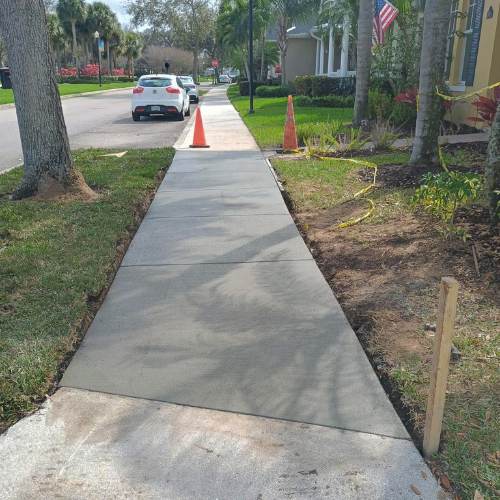 Newly poured concrete sidewalk with orange traffic cones, next to grass and street.