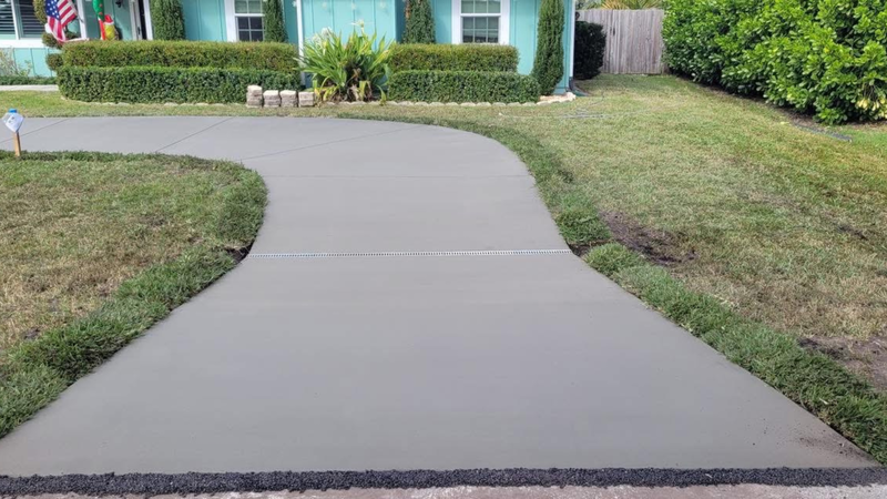 Curving concrete driveway leads to a light blue house with green landscaping.