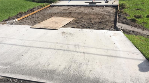 Concrete driveway with a section of dirt preparation. A piece of plywood lies on the dirt.