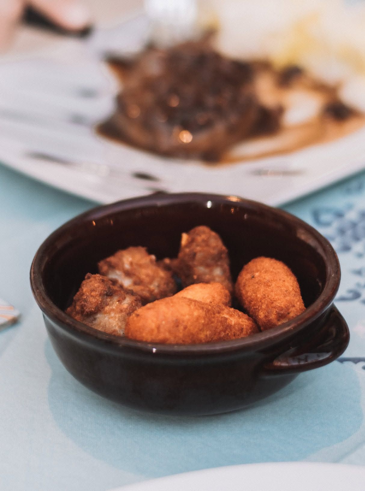 Ciotolina marrone con stuzzichini fritti su un tavolo azzurro chiaro, con un piatto sfocato sullo sfondo