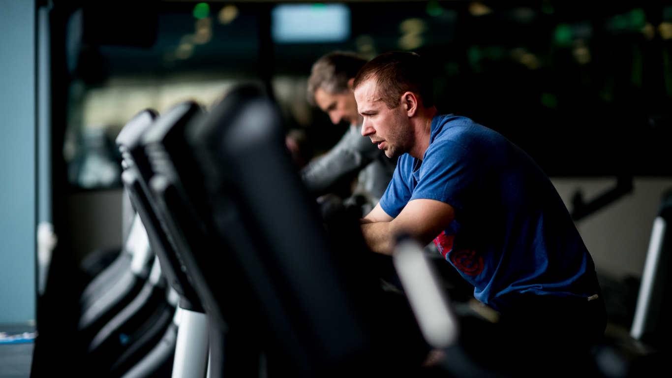 Two men exercising on stationary bikes in a gym, with the foreground rider in a blue shirt.