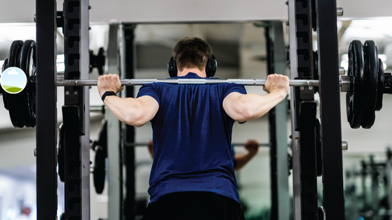 Person lifting a barbell on a squat rack in a gym, seen from behind, wearing a blue shirt.