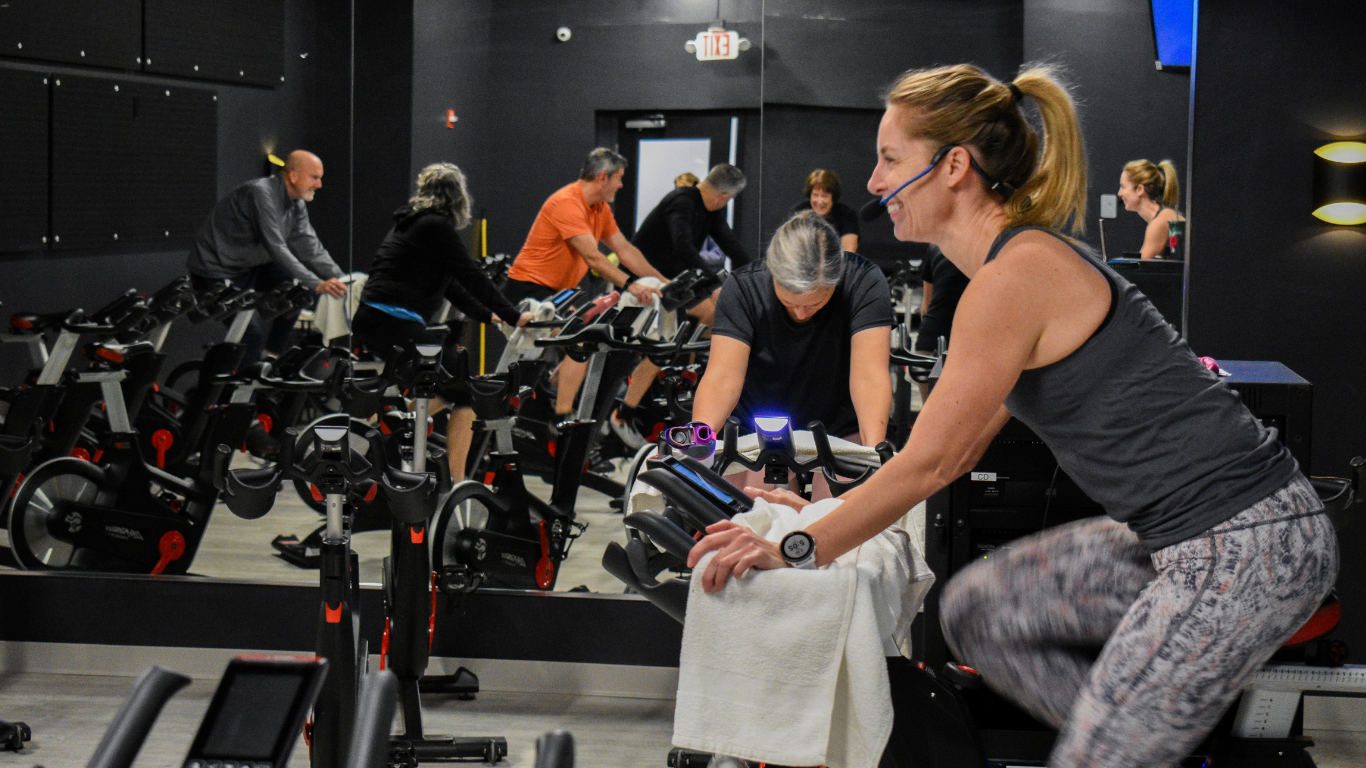 Cycling studio class with riders on stationary bikes in front of wall mirrors; woman in foreground pedaling.