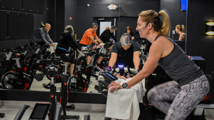 People exercising on stationary bikes in a dim indoor studio, with a woman in the foreground smiling.