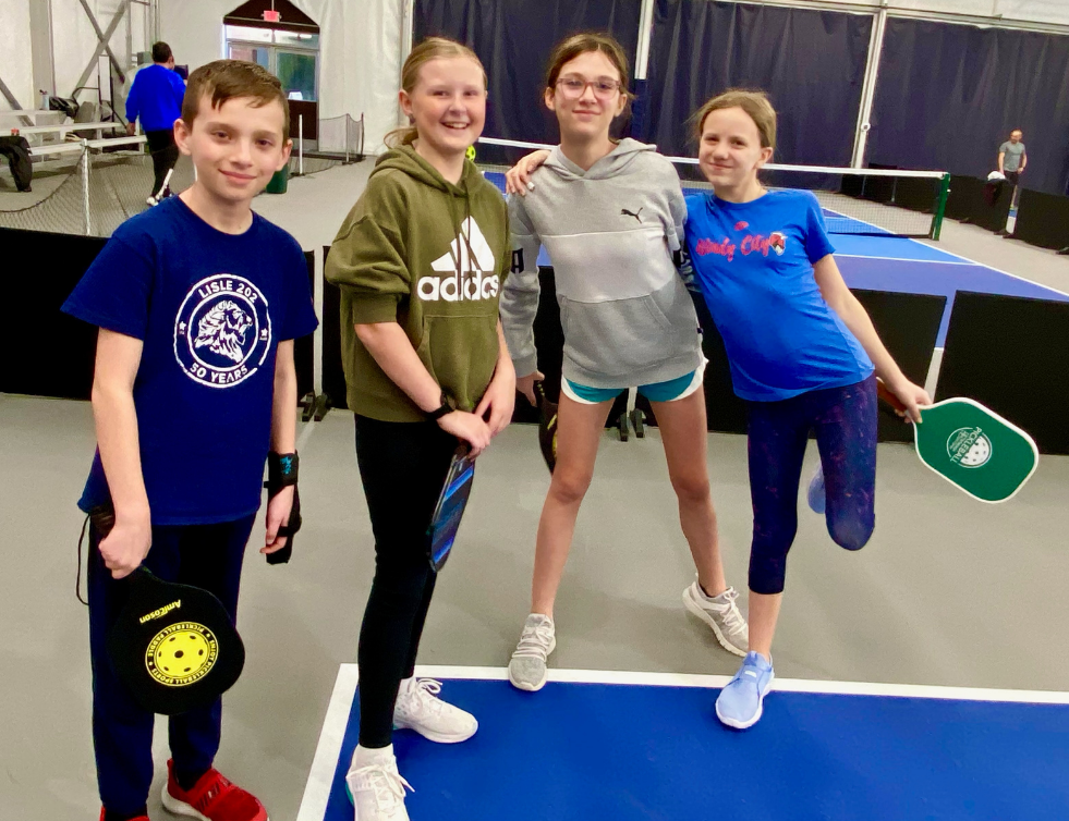 Four people holding pickleball paddles pose smiling on an indoor court.