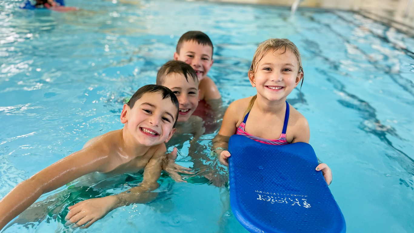 Three children smiling in a swimming pool, one holding a blue kickboard.