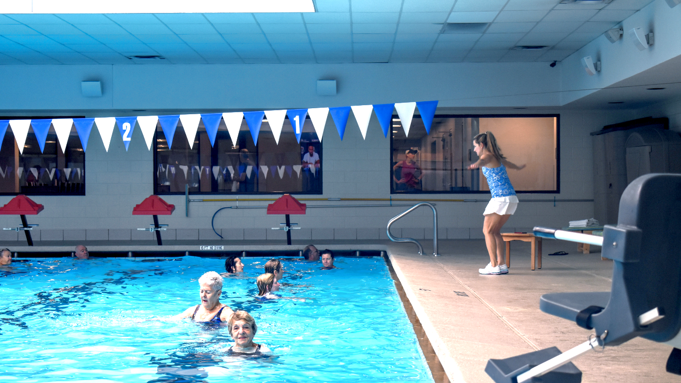 Indoor pool with swimmers and a woman standing on the deck near a diving board.
