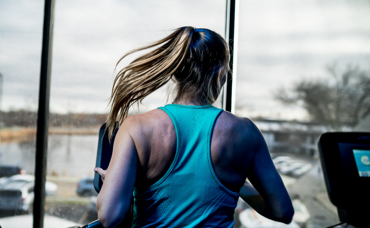 Woman running on a treadmill in a gym, wearing a teal tank top with a ponytail swinging.