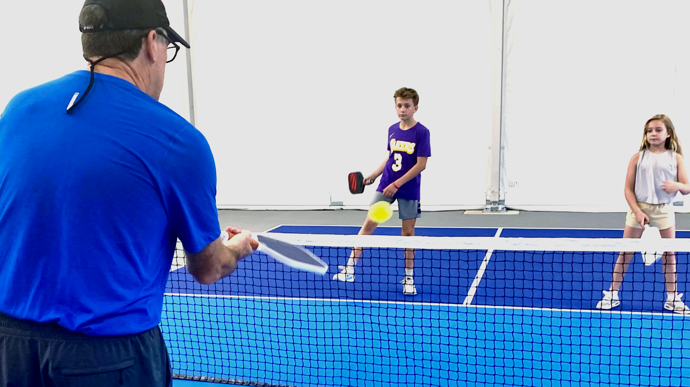 Two players rally at an indoor pickleball net while a man in blue watches from the foreground.
