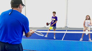 Man in blue shirt playing pickleball with two players across the net indoors