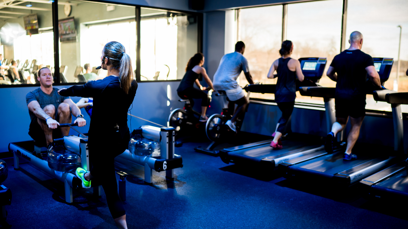 People exercising on rowing machines and treadmills in a bright gym.