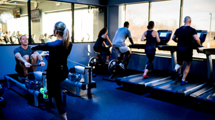 People exercising in a gym on treadmills and stationary bikes, with bright windows in the background