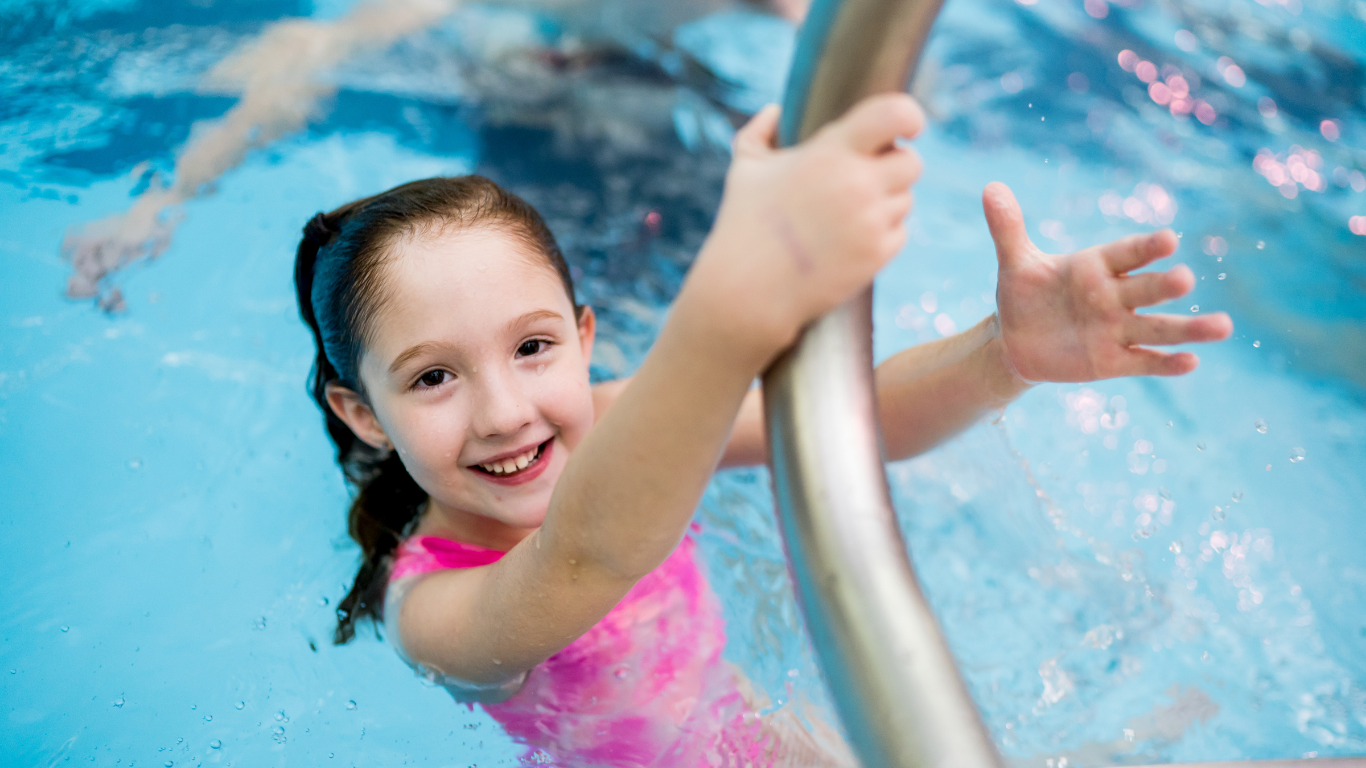 Smiling child in a pink swimsuit holding a pool ladder in a blue swimming pool