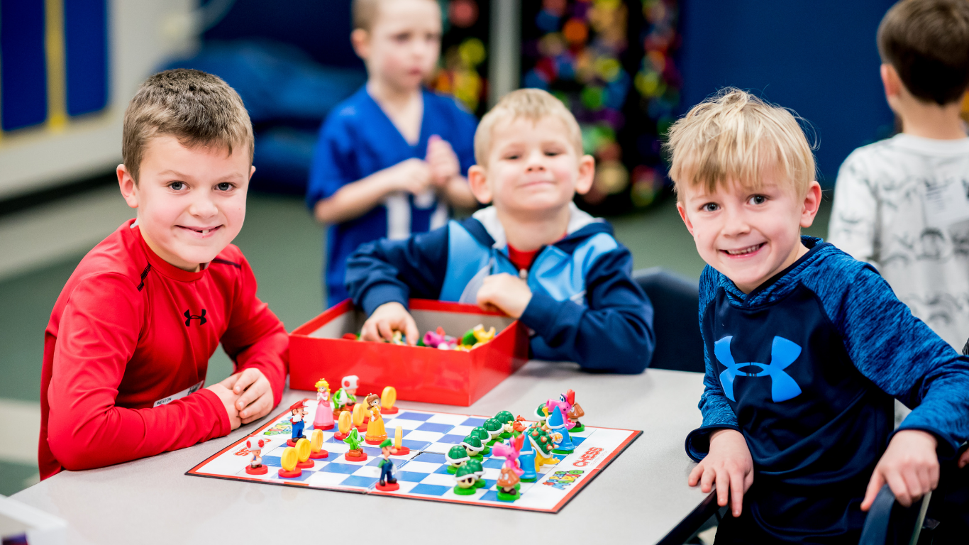 Children playing a colorful board game at a table, with two smiling kids in the foreground.