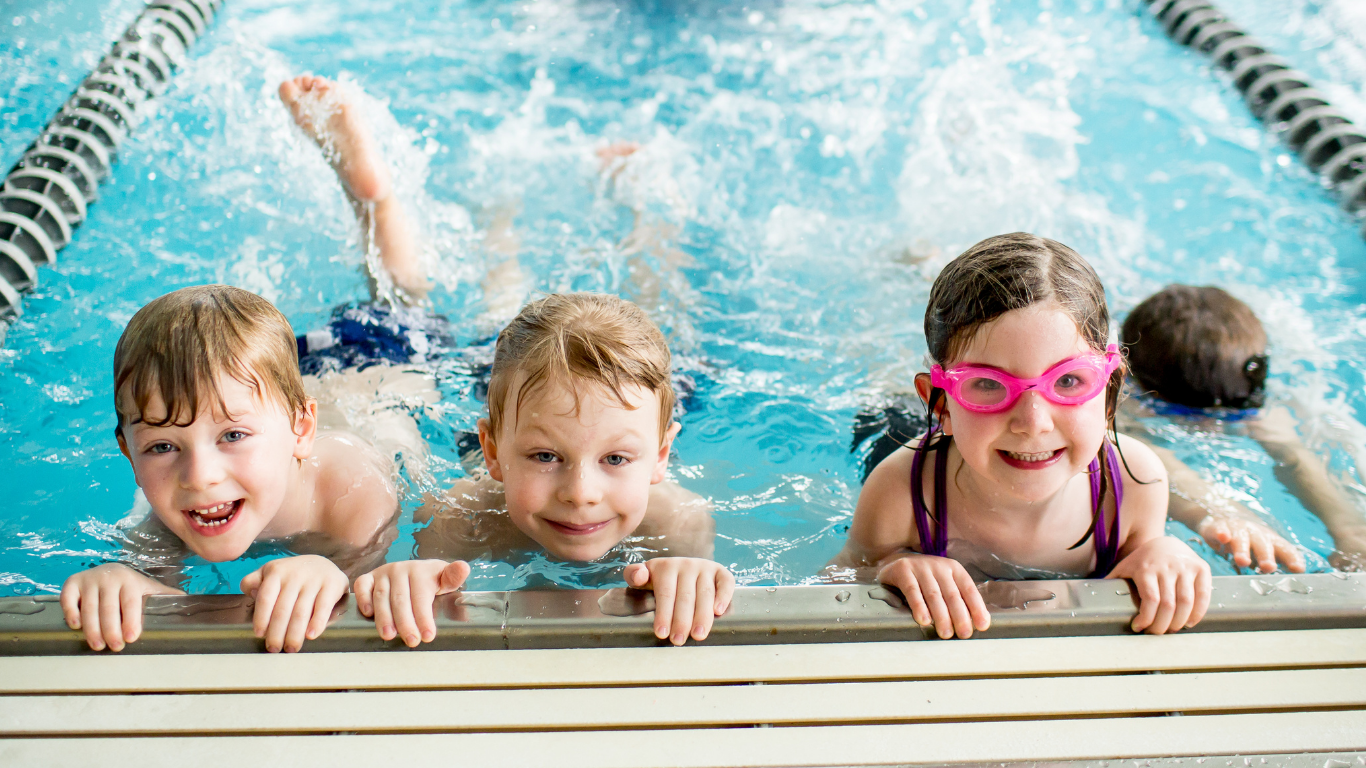 Three children holding pool edge, smiling in a swimming pool, with one wearing pink goggles.