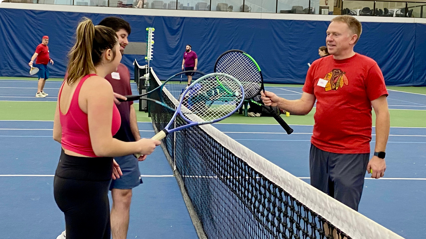 People at an indoor tennis net discussing rackets on a blue court.