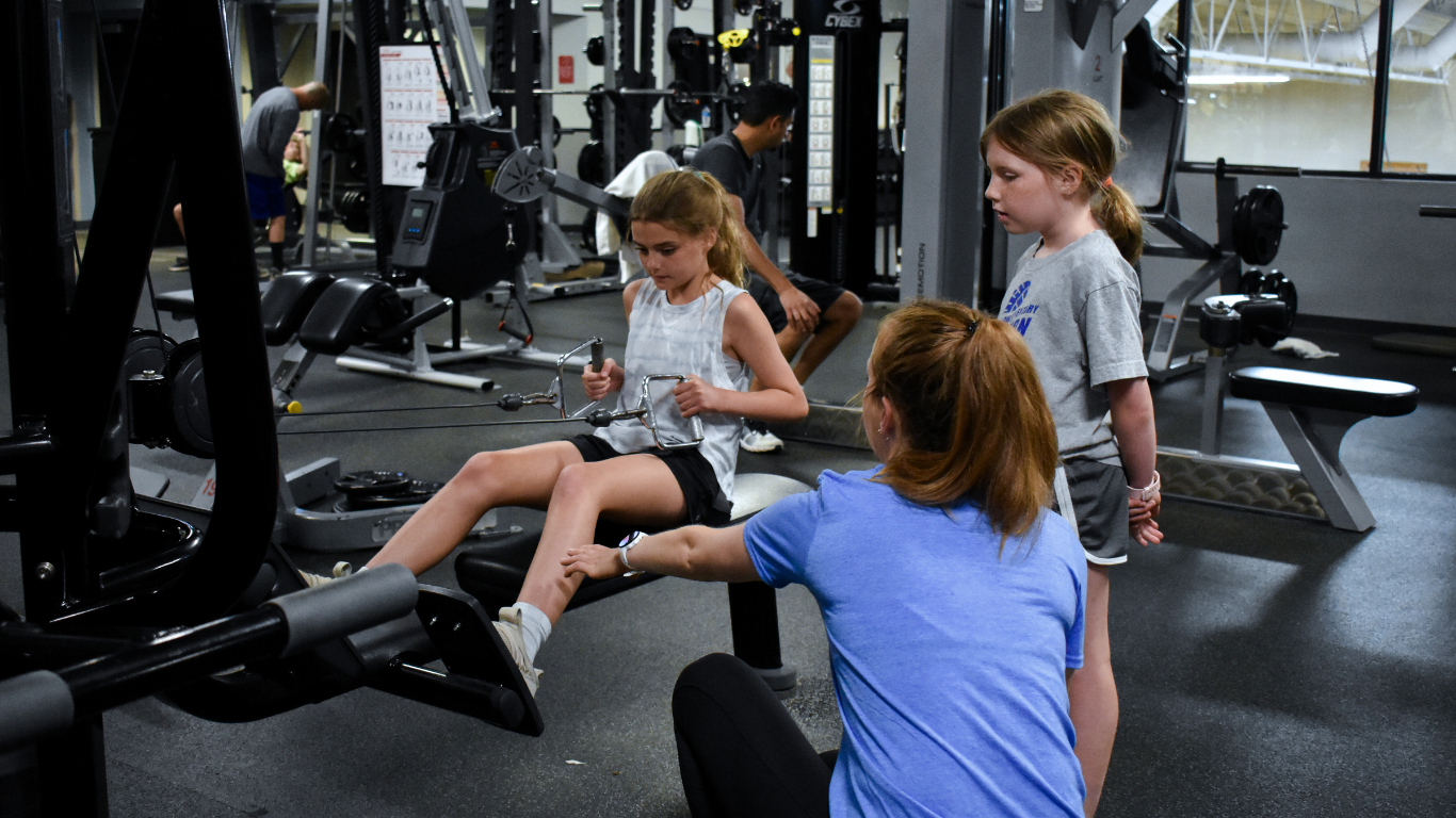 Children using gym equipment while an adult supervises in a fitness center
