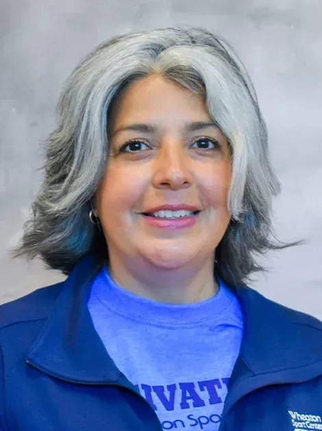 Smiling person with gray hair wearing a blue jacket and purple shirt against a plain gray background