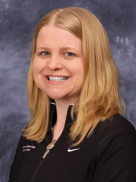 Portrait of a smiling person with shoulder-length blonde hair in a black Nike jacket against a gray backdrop