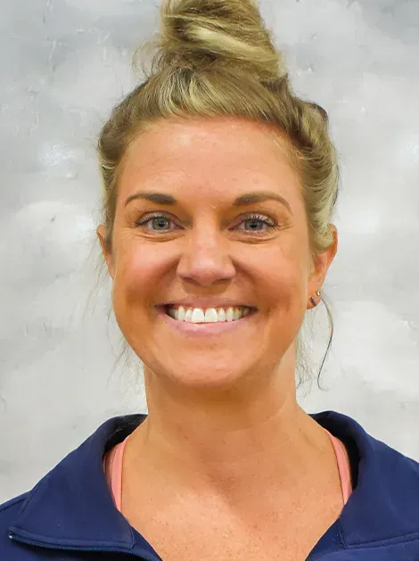 Smiling woman with blonde hair in a top bun wearing a navy jacket against a gray background