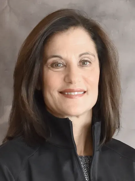 Portrait of a woman with dark hair wearing a black jacket, smiling against a gray backdrop