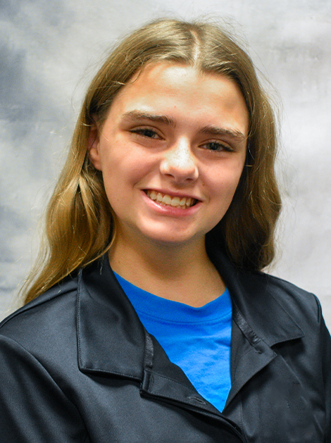 Smiling person with shoulder-length hair wearing a black jacket over a blue shirt against a gray background