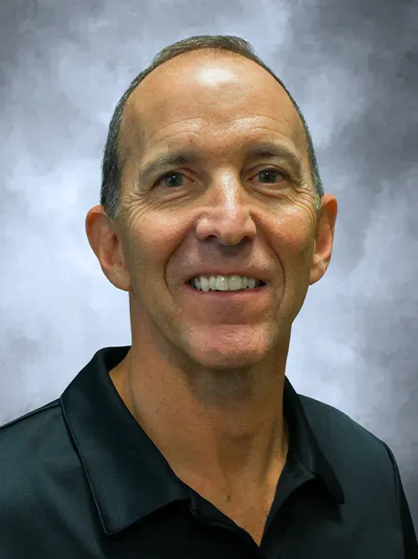 Head-and-shoulders portrait of a smiling man in a dark polo shirt against a blue-gray background