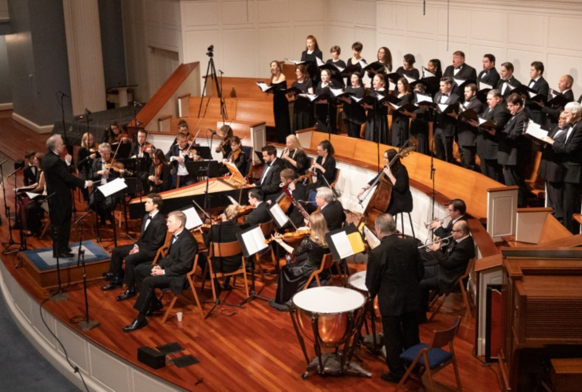 Choir and orchestra performing on a concert hall stage with wooden floors and black formal attire