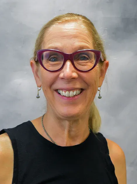 Smiling person wearing purple glasses and a black sleeveless top against a gray background