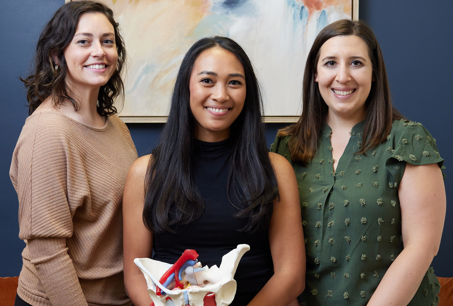 Three smiling women standing together, holding a small object, in front of a colorful abstract painting.