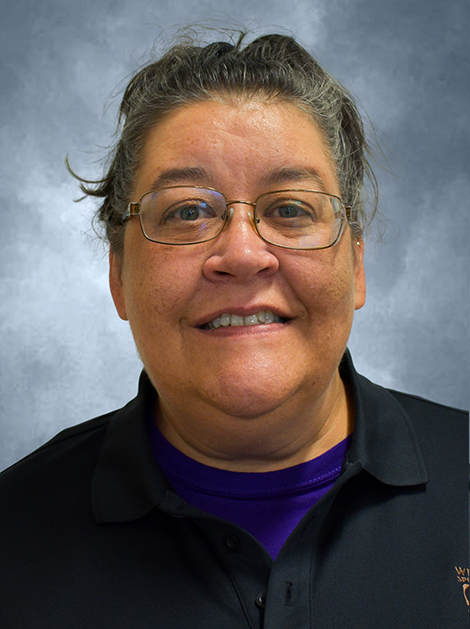 Head-and-shoulders portrait of a smiling person in glasses, wearing a dark shirt and purple top against a gray background