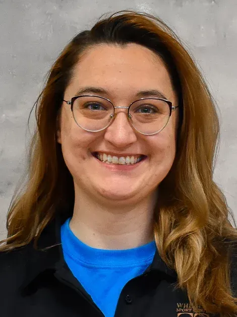 Smiling person with glasses and brown hair wearing a black shirt over a bright blue top against a gray background
