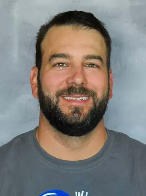 Smiling man with dark hair and beard wearing a gray shirt against a plain gray backdrop
