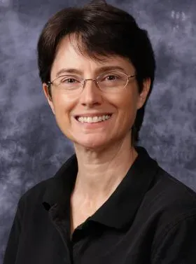 Portrait of a smiling person with short dark hair and glasses, wearing a black collared shirt against a gray backdrop