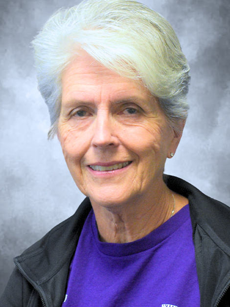 Portrait of a smiling person with short white hair, wearing a purple shirt and dark jacket against a gray background