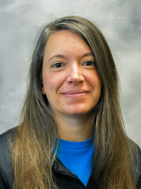 Headshot of a person with long brown hair, wearing a blue shirt and black jacket, against a gray background