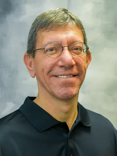 Portrait of a smiling man in glasses wearing a dark polo shirt against a gray backdrop