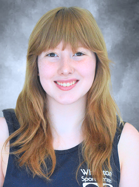 Smiling person with long strawberry-blonde hair in a dark sleeveless top against a gray backdrop