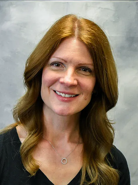 Smiling woman with long reddish hair wearing a black top against a gray background