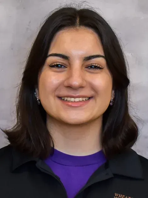 Smiling person with dark hair wearing a black jacket over a purple shirt against a gray background
