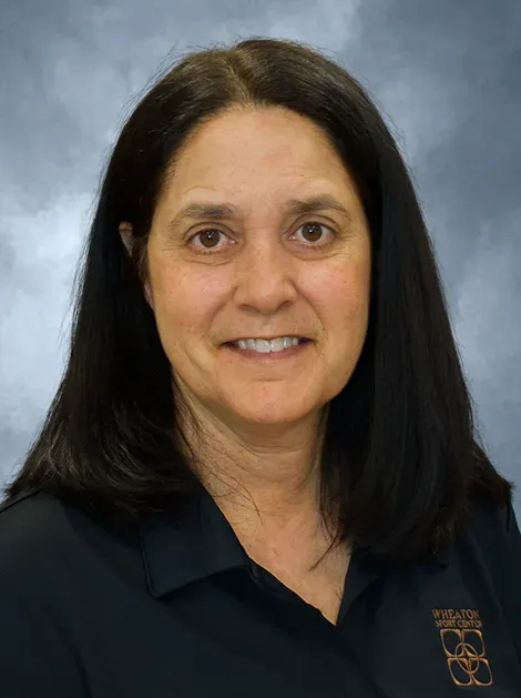 Portrait of a smiling person with long dark hair, wearing a dark polo shirt against a blue-gray backdrop