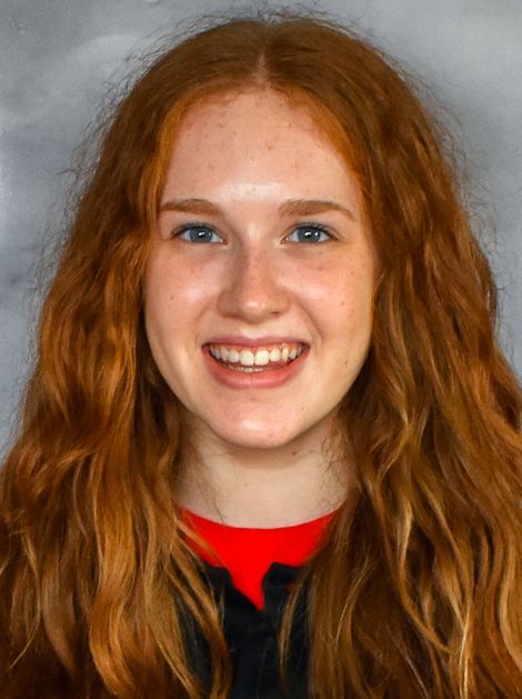 Smiling person with long red hair wearing a dark top and red shirt against a gray background