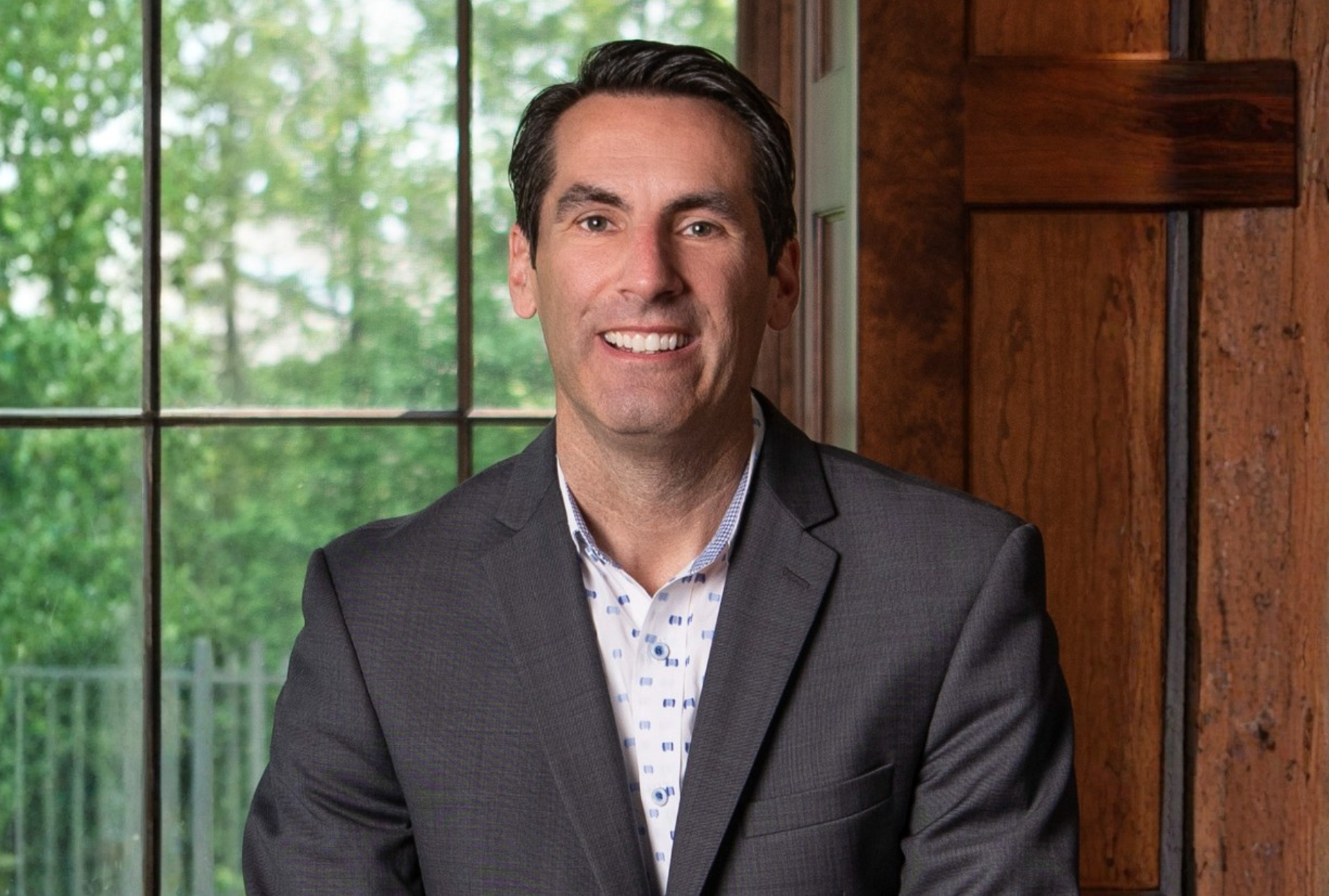 Smiling man in a gray suit seated indoors beside a window and wooden wall