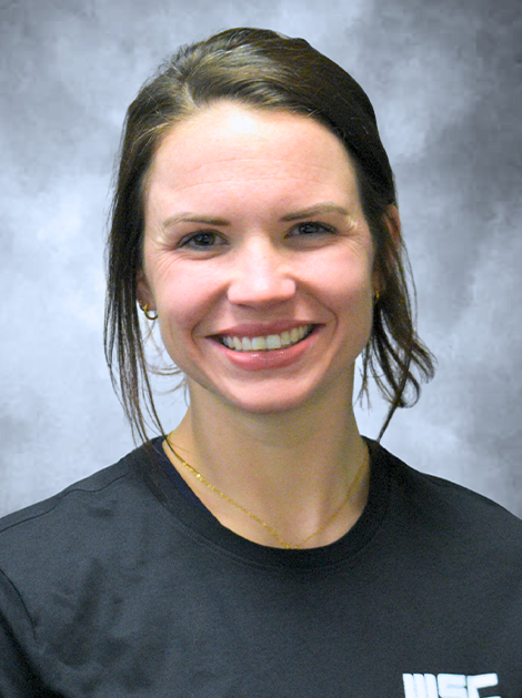 Smiling person in a dark shirt against a gray studio backdrop, wearing a white MEC logo