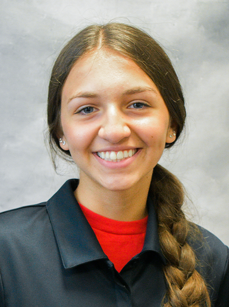 Smiling person in a dark jacket with a red shirt and braided hair against a gray background