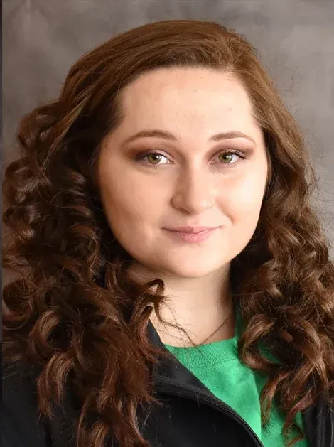 Portrait of a smiling woman with curly brown hair, wearing a black jacket and green top against a neutral background